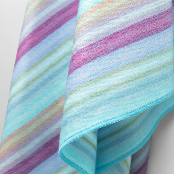 Tzacantza baby alpaca wool blanket – a colorful, diagonally striped fabric hangs, showcasing shades of blue, pink, purple, and green. The fabric is draped against a simple, plain background