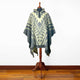 A poncho with geometric patterns is displayed on a wooden mannequin in a room with wooden flooring and a plain white wall background.