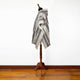 A striped poncho with a hood drapes on a wooden mannequin stand, set in a room with polished wooden flooring and a plain white wall background.