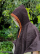 A person wearing a brown hooded cloak with a red, patterned trim stands amidst lush green foliage, partially obscured in profile, creating a natural and mysterious atmosphere.
