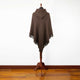 A brown poncho with fringe trim is displayed on a wooden stand in a room with a polished wooden floor and plain white wall.