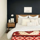 A neatly made bed with patterned pillows and a colorful geometric blanket sits against a dark accent wall. A wooden nightstand with a lamp and decor completes the modern bedroom setting.