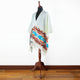 A white poncho with colorful geometric patterns is draped over a mannequin. It features blue, red, and brown designs with tassels, set against a wooden floor and a plain background.