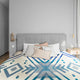 A bed is adorned with multiple pillows and a cream and blue geometric-patterned blanket. It stands against a grey upholstered headboard, flanked by a simple nightstand and a wall lamp.