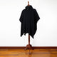 Achema llama wool hooded poncho – a black poncho drapes over a wooden mannequin stand. It features a high collar with a button. The scene is set against a plain white wall and wooden floor