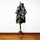 A black and white patterned poncho drapes over a wooden mannequin stand, placed in a room with a polished wooden floor and a plain white wall backdrop.