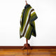 A striped poncho with green, white, and black hues drapes over a wooden mannequin stand, placed in a room with wooden flooring and a plain white wall.