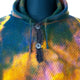 A multi-colored knitted hoodie features a buttoned neckline. The colors blend in a tie-dye pattern, primarily green, yellow, and blue, creating a vibrant effect on a mannequin display.