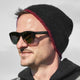 A person wears a black beanie and sunglasses with HAWKERS on the side, standing outdoors in natural light.