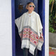A person stands in a doorway, wearing a cream-colored poncho with geometric patterns, sunglasses, and striped pants. The dark blue door behind contrasts with the white wall and greenery outside.
