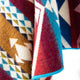 A colorful, patterned wool blanket featuring geometric shapes in red, blue, yellow, and white hangs with visible texture and detail, against a plain white background.