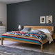 A wooden bed features a colorful geometric-patterned blanket, surrounded by white pillows. Positioned in a modern bedroom, it includes a dark wall, abstract art, and a dresser with a vase.
