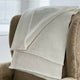 A cream-colored knitted blanket is draped over the arm of a patterned beige chair, situated in a softly lit room with large windows in the background.