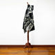 A black poncho with white geometric patterns is displayed on a wooden stand in a room with a wooden floor and a plain white wall.