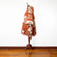 A mannequin is draped in a geometric-patterned, orange and cream poncho. It stands in a minimalist room with a hardwood floor and a plain white wall backdrop.