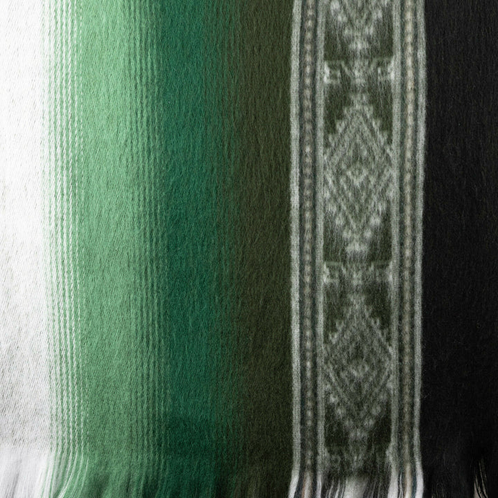 A woven fabric displays a gradient of green and black stripes, featuring intricate geometric patterns on the right, with fringed edges at the bottom.