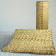 A woven straw mat lies flat, with a matching rolled-up mat standing beside it, placed on a light wooden floor in a minimalistic room.