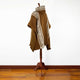 A brown poncho with geometric patterns hangs on a wooden stand against a plain white wall, situated on a polished hardwood floor.