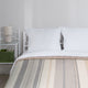 Yankuam baby alpaca wool blanket – a bed with white pillows is covered by a striped blanket. Beside it, a nightstand holds a small lamp and basket with books, against a plain white wall