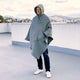 A person stands on a rooftop wearing a gray hooded poncho, with one hand in a pocket. In the background, a cityscape and mountains are visible under a cloudy sky.