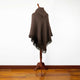 A brown fringed poncho drapes over a wooden mannequin stand on a wooden floor, set against a plain white background.