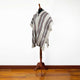 A striped poncho hangs on a wooden mannequin stand in a room with wooden flooring, against a plain white wall.