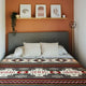Bed with patterned blanket, adorned with white pillows, sits against a sepia-toned wall. Above, a shelf displays framed art and a small plant, accompanied by a geometric floor lamp.
