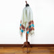 A white poncho with colorful, geometric patterns hangs on a wooden stand in a room with a wooden floor and a white wall.