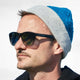 A person wearing HAWKERS sunglasses and a blue and gray beanie gazes sideways in a sunlit setting.