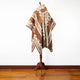 A brown and beige patterned poncho with geometric designs is draped on a wooden mannequin stand. It is set against a plain white wall on a hardwood floor.