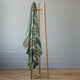 Jambue baby alpaca wool blanket – a striped, multicolored blanket hangs from a minimalist, wooden coat rack in a room with light gray walls and a wooden floor