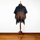 A black poncho with geometric patterns and fringed edges is displayed on a wooden mannequin stand in a minimalist room with a hardwood floor.