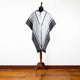 Poncho hangs on a wooden stand, featuring a hood, two buttons, and a geometric design. The poncho's colors are gray and white. It is displayed indoors on a wooden floor.