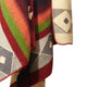 A multicolored woven blanket drapes, showcasing geometric patterns in red, green, orange, and cream. The design features diamond shapes and lines, with visible texture in a close-up view.
