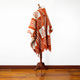 A patterned, rust-colored poncho with a geometric design is displayed on a wooden stand. It’s draped elegantly in a well-lit room with a wooden floor and white walls.