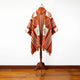 Poncho on a mannequin, showcasing red and white geometric patterns, hangs motionless in a room with wooden floors and a plain white wall backdrop.