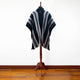 A black poncho with gray and red diagonal stripes is displayed on a wooden mannequin stand in a room with wooden flooring and a plain white wall.