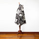 A patterned poncho drapes over a wooden mannequin stand, showcasing geometric designs. Positioned against a white wall, it rests on a polished wooden floor.