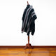 A striped poncho drapes elegantly over a wooden mannequin stand. The poncho features black, gray, and maroon stripes, set in a room with a polished wood floor and white walls.