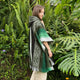 A person wears a green and gray patterned poncho, standing among lush green ferns and tropical plants, surrounded by vibrant foliage and a serene outdoor atmosphere.