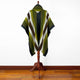 A poncho with green, black, and white chevron stripes is displayed on a wooden mannequin stand, set against a plain white wall and hardwood floor.