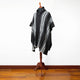 A striped poncho is displayed on a wooden mannequin stand. The poncho features black and white vertical stripes. The background is a plain white wall with a wooden floor.