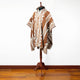 A poncho with geometric patterns and earthy tones drapes over a wooden mannequin stand, set on a polished hardwood floor against a plain white background.