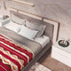 Bed with gray pillows and red geometric-patterned blanket lies in a modern bedroom. A textured wall features a light fixture above, and a nightstand holds books and cosmetics.