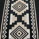 A woven fabric features repeating geometric patterns with diamond shapes and zigzag borders, in black and beige colors, creating a symmetrical design.