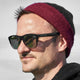 A person wears black HAWKERS sunglasses and a maroon knit beanie against a blurred outdoor background, highlighting a sunny day with clear skies.