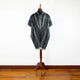 Nambija lightweight alpaca wool hooded poncho – a mannequin displays a black and gray patterned poncho with fringed edges, standing on a wooden floor against a plain white background