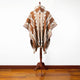 A poncho with brown, beige, and white geometric patterns hangs on a wooden stand in a room with a white wall and wooden floor.