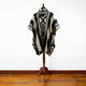 A poncho with geometric patterns is displayed on a wooden mannequin stand. The background is a plain white wall with a wooden floor.