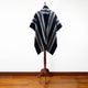 A striped poncho drapes over a wooden stand. It features black, gray, and burgundy diagonal stripes. The setting is a room with a polished wooden floor and a plain white wall.